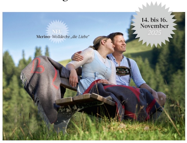 Ein Paar in Tracht sitzt auf einer Bank vor Wald und blauem Himmel. Über der Lehne liegt eine Schafwolldecke.